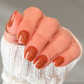 warm orange glitter nail wraps, with a white knit sleeve and a thin gold ring against a soft gray background.